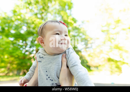 Baby girl being held up outdoors in front of tree looking to the right