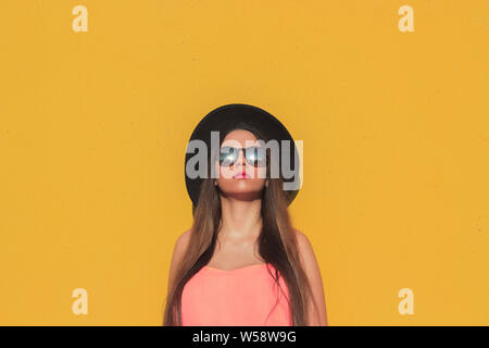Girl standing wearing fashionable sunglasses and black hat with a yellow wall as background Stock Photo