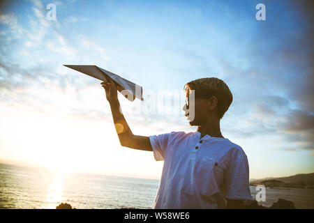 Happy child playing with paper airplane at sunset, dreaming about flying, Plane in teen hand and blue sky with clouds and sun. Boy have fun during sun Stock Photo