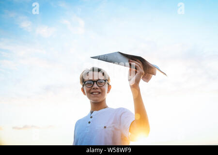 Happy child playing with paper airplane at sunset, dreaming about flying, Plane in teen hand and blue sky with clouds and sun. Boy have fun during sun Stock Photo