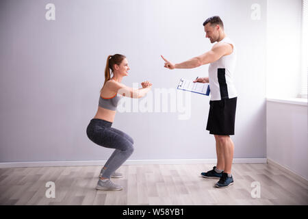 Side View Of Young Sporty Woman Doing Deep Squat Exercise In Front Of Gym Trainer Stock Photo