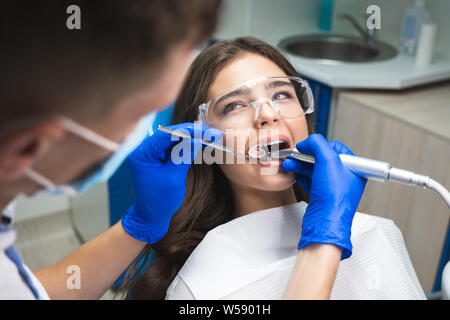 dentist in mask filling the patient's root canal while she is lying on dental chair in safety glasses under the medical lamp in clinic Stock Photo