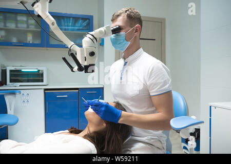 handsome dentist in medical mask examining patient woman laying on the dental chair using microscope in clinic Stock Photo