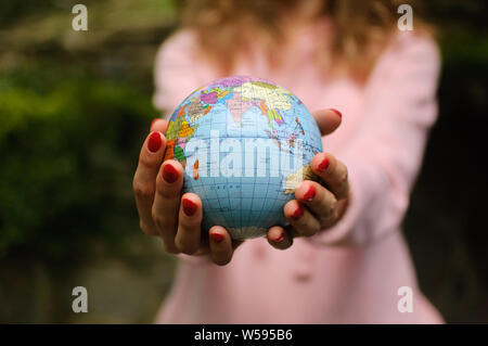 Young female teenager in pink casual clothes with red manicure holding a little globe with geografical names in Ukrainian cyrillic letters on it in Stock Photo