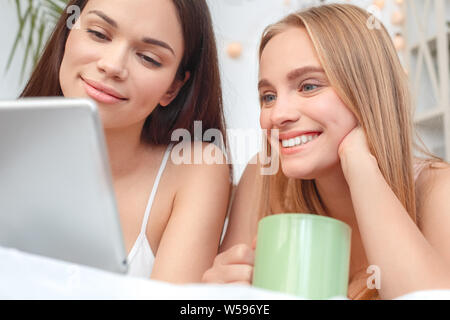 Young women rest together at home lying on bed drinking hot coffee watching video online on digital tablet smiling joyful close-up Stock Photo