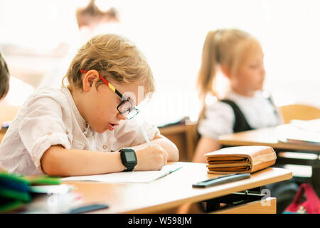 Blonde boy with big black glasses sitting in classroom, studing, smiling. Education on elementary school, first day at school. Stock Photo