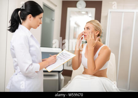 Facial care. Serious young woman touching her face and telling a beauty doctor in front of her about needed procedures. Stock Photo