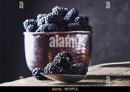 Ripe blackberries in old copper dishes on old wooden boards and lying next to a spoon on a dark background. Stock Photo