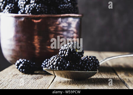 Ripe blackberries in old copper dishes on old wooden boards and lying next to a spoon on a dark background. Stock Photo