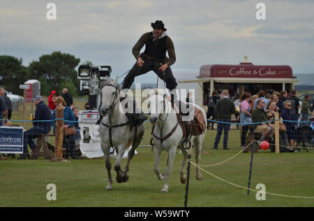 A man in Cowboy dress riding astride on two horses as part of a Wild West horse display at the 2019 Sutherland County Agricultural Show, Scotland, UK Stock Photo