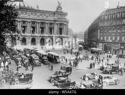 An early 20th century black and white photograph taken in Paris, France. The main building is the Acadamie National de Musique, also known as the Opera de Paris, The Royal Academy of Music, and the Palais Garner. The photo shows a busy scene, with many people, cars and buses. Stock Photo