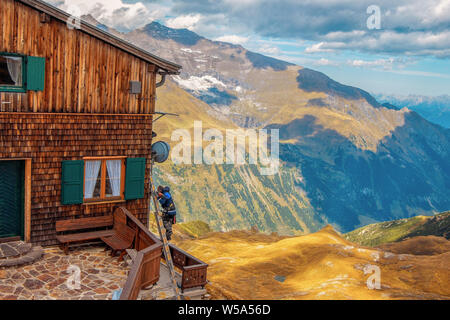 A worker with tools is standing on the stairs by a wooden house in the mountains against the backdrop of a magnificent autumn landscape. Stock Photo