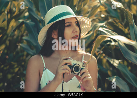 Outdoor portrait of a young hispanic girl wearing summer dress and hat taking photos Stock Photo