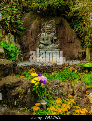 Small statue with rich detailed artwork placed on rocks surrounded by flowers in Kamakuras Hase-dera temple, Japan November 2018 Stock Photo