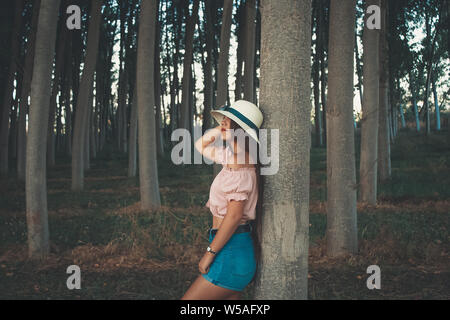 Young long haired woman leaning on a tree wearing white sun hat short blue jeans and pink blouse Stock Photo