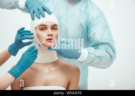 Stop aging. Young attractive woman with head in bandages looking at camera while doctors making plastic surgery operation. Plastic surgeons in blue gloves holding scalpel and syringe near patient face. Facelifting. Cosmetic procedures Stock Photo