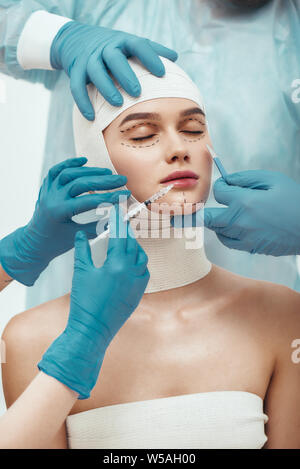 No chance for wrinkles. Young attractive woman with head in bandages keeping eyes closed while plastic surgeons in blue gloves are working with her face. Doctors holding scalpel and syringe near patient face. Facelifting. Stop aging. Beauty. Stock Photo
