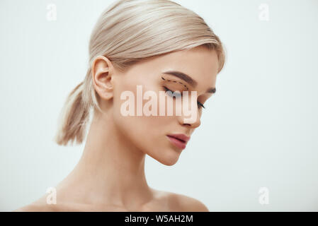 Before facial surgery. Side view of pretty young blonde woman with sketch on her face standing against grey background. Plastic surgery concept. Healthcare. Beauty concept. Stock Photo