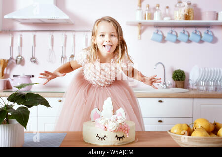 Little girl stands near the table with a birthday cake and shows tongue. Stock Photo