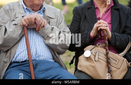 Senior couple holding hands while sitting on the park bench together. Retired man and woman with walking cane.  Close up of hands. Stock Photo