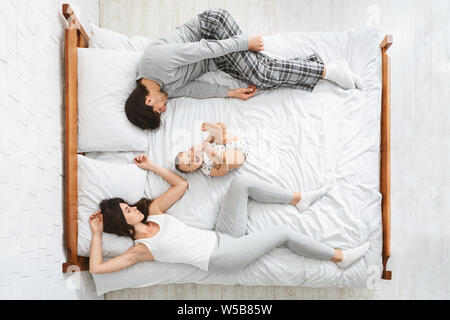Exhausted parents sleeping on sides of bed, active baby playing in middle Stock Photo