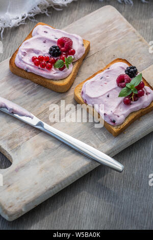 Toast with mixed berries on wipped cream on a wood board on light background. Summer traditional homemade dessert.