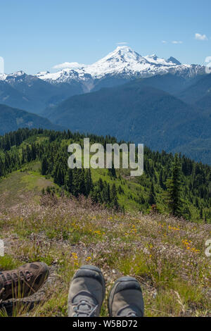 Looking across alpine meadows and forested ridges from High Divide Trail towards snow-covered Mt Baker... the perfect spot for hikers to sit down and Stock Photo