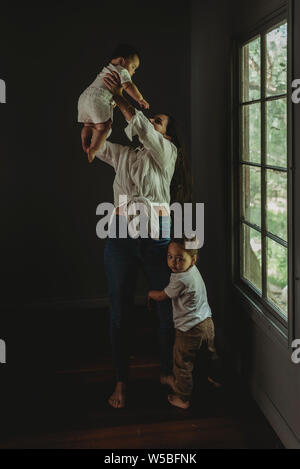 Young mother lifting baby daughter in the air while son hugs her Stock Photo