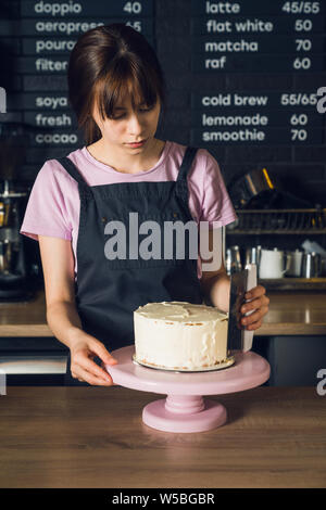 Young woman in dark apron and pink t-shirt smoothing cream on a side of cake by spatula in the cafeteria. Stock Photo