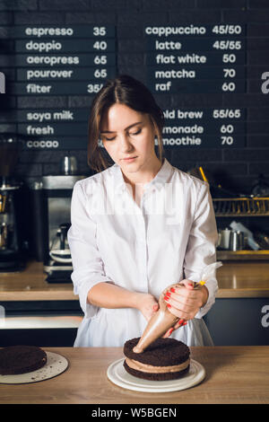 Young and beautiful woman in white shirt squeezes out cream on cake in Stock Photo