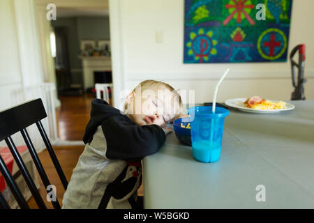 Tired toddler boy falls asleep while sitting in dining room chair Stock Photo