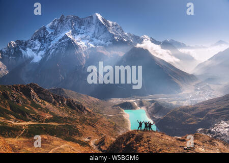Happy people with raised up arms on the mountain peak against beautiful lake, snowy mountain at sunrise. Landscape with man and woman, rocks in clouds Stock Photo