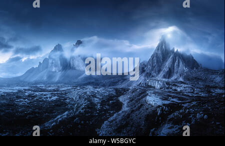 Mountains in fog at beautiful night. Dreamy landscape with mountain peaks, stones, grass, blue sky with blurred low clouds, stars and moon. Rocks at d Stock Photo