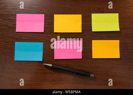 Six colorful sticky notes in two row and pen on brown wooden desk. Top view and space for text. Close up, selective focus Stock Photo