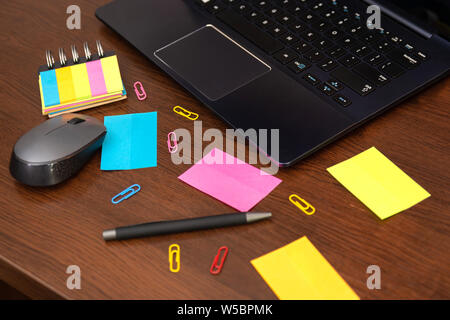 Colorful sticky notes and spilled paper clips, pen, computer mouse and laptop on brown wooden desk. Top view and space for text. Close up Stock Photo