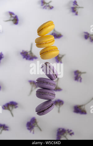 Traditional French, colorful, purple and yellow macaroons cookies in motion falling (levitation) on a white with flowers background. Stock Photo