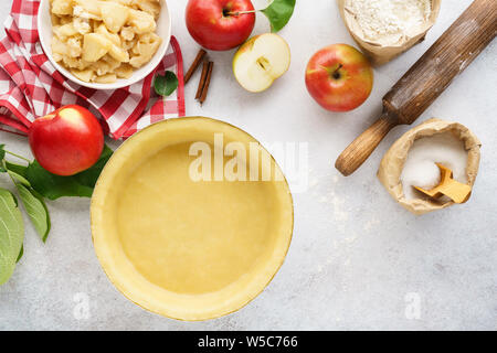 Rolled pie crust in a baking dish with filling, tools and ingredients. Apple pie making process. Copy space for recipe. Stock Photo