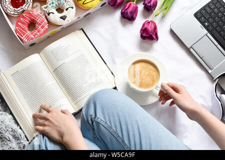 Girl reading a book and has brealfast in bed. Tray with strawberry pink donut with coffee and flowers. Doughnuts from food delivery. Top view. Stock Photo