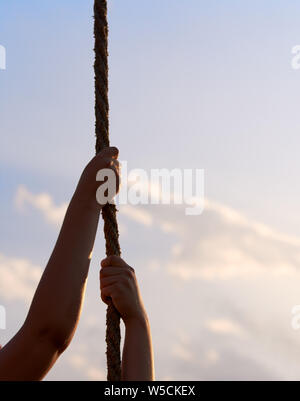 Young woman or girl holds hands on rope and climbs up in physical education class. Blue sky with clouds. Stock Photo