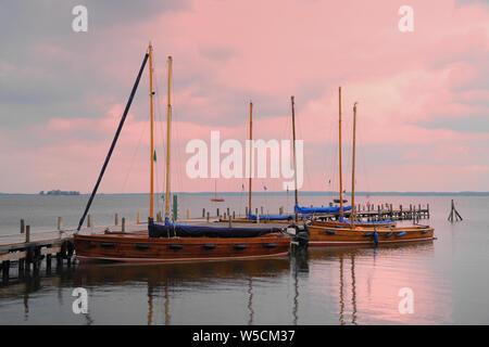 Wooden sailboats moored to a jetty on a calm lake against a colorful pink sky. Stock Photo