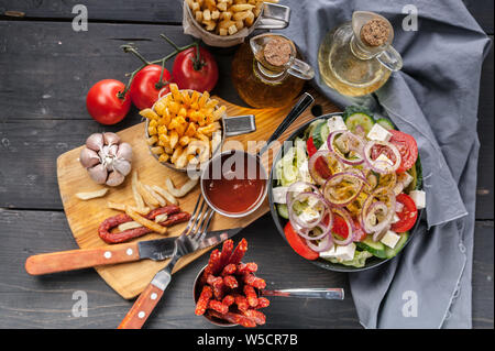 Homemade French Fries and Greek Salad. Onions, garlic, smoked sausages, tomato sauce, fork and spoon on a dark wooden table. Top view Stock Photo