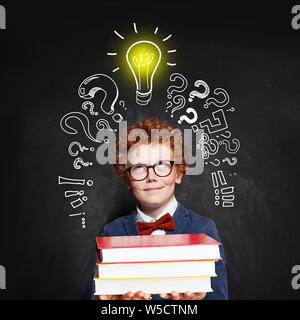 Little boy wearing bow tie and suit with lightbulb and books on blackboard background. Brainstorming and idea concept Stock Photo