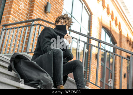 Young attractive pensive man sitting on concrete steps of modern red bricked building and wrapping up in black coat. Stock Photo