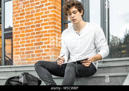 Attractive man holding notepad in his hands and sitting on concrete steps of red bricked building. Stock Photo