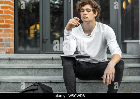 Attractive pensive student sitting on modern building steps and making notes in his black notepad. Stock Photo