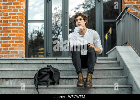 Attractive pensive student sitting on modern building steps and making notes in his black notepad. Stock Photo