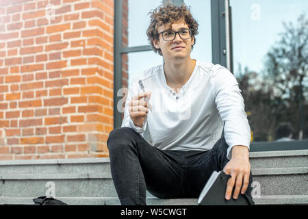 Pensive young attractive man sitting on concrete steps of modern building and holding notepad in hands while doing his homework. Stock Photo