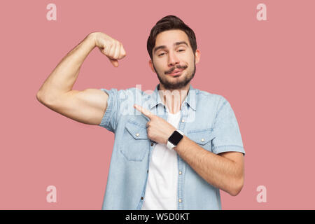 I am strong and I can do anything. Portrait of proud handsome bearded young man in blue casual shirt standing pointing and showing his strong bicep. i Stock Photo