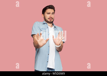 Portrait of scared bearded young man in blue casual style shirt standing with afraid or worry face, looking at camera and blocking with his hands. ind Stock Photo