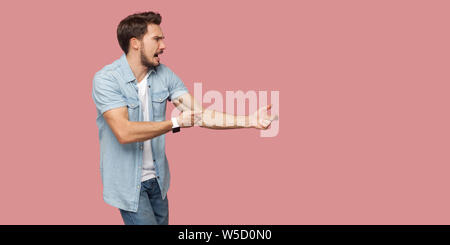 Profile side view portrait of serious screaming bearded young man in blue casual shirt standing in attack or pulling hands gesture and looking forward Stock Photo